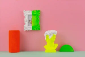 Eclectic space styling with vibrant yellow bust sculpture, neon green and white ornate frame, and colorful décor against a pink wall.