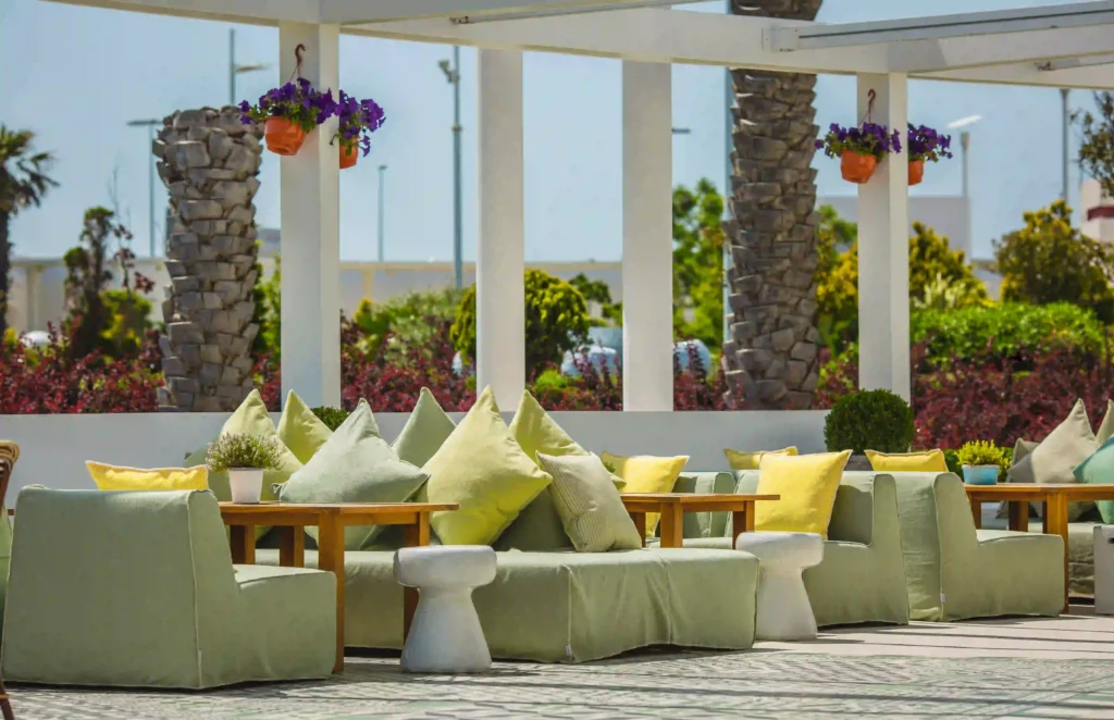 Modern outdoor terrace seating with green and yellow cushions, wooden tables, and hanging flower pots under a white pergola on a sunny day.