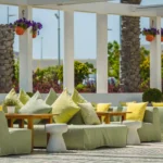 Modern outdoor terrace seating with green and yellow cushions, wooden tables, and hanging flower pots under a white pergola on a sunny day.