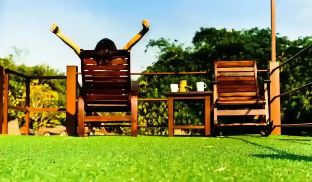 A person sitting on a wooden chair outdoors, stretching their arms up toward the sky, with green artificial grass in the foreground, a wooden table with cups in the middle, and trees in the background under a clear blue sky