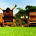 A person sitting on a wooden chair outdoors, stretching their arms up toward the sky, with green artificial grass in the foreground, a wooden table with cups in the middle, and trees in the background under a clear blue sky