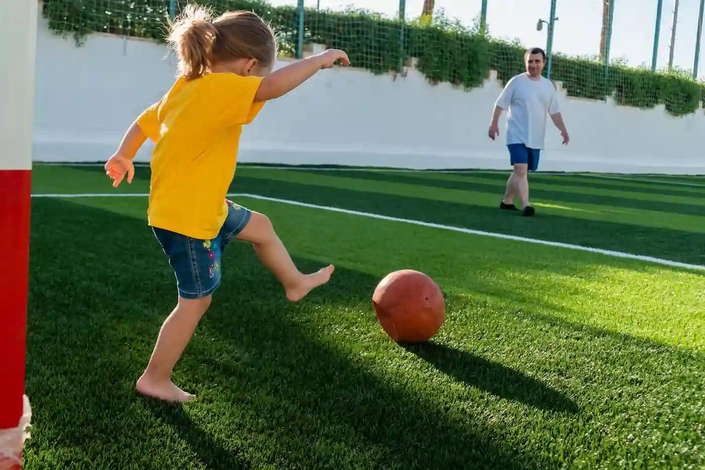 A young child in a yellow shirt and denim shorts kicks a ball barefoot on a green artificial turf field, while an adult watches in the background.