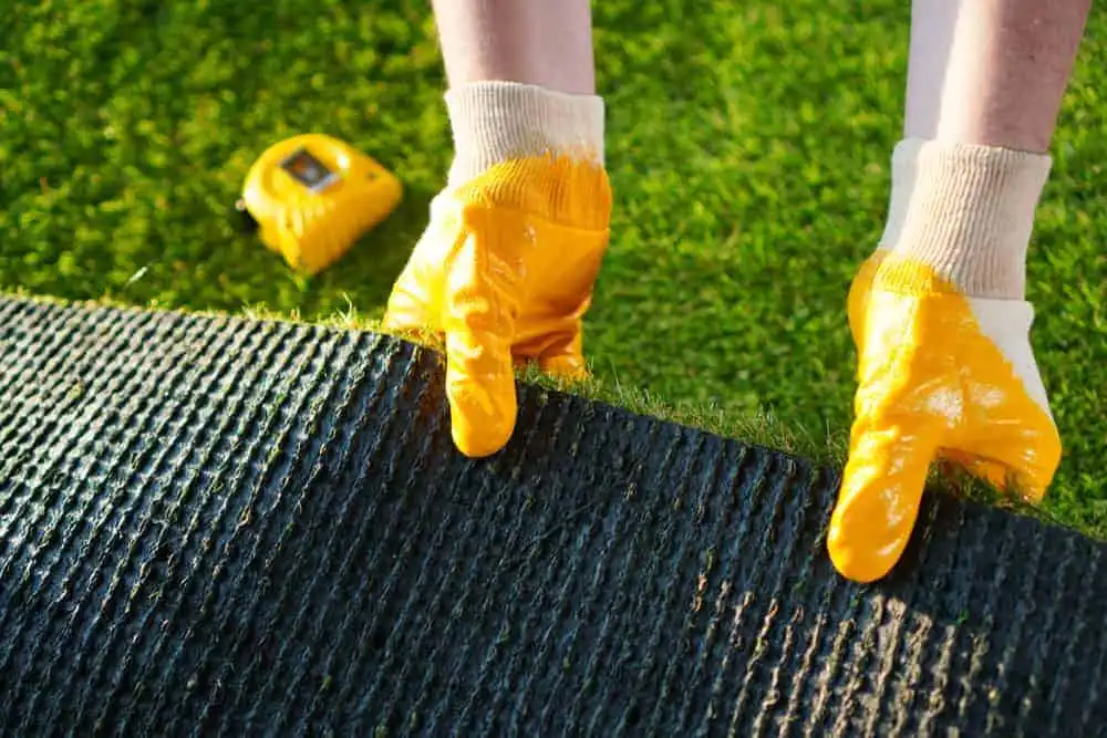 Worker wearing yellow gloves installing artificial grass with measuring tape in the background.