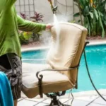 Person cleaning outdoor chair cushion with hose near swimming pool