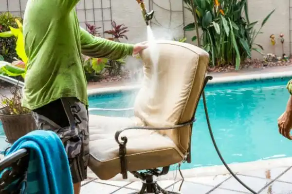 Person cleaning outdoor chair cushion with hose near swimming pool