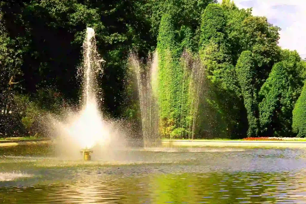 Outdoor water fountain spraying high jets of water in a pond with lush green trees in the background.