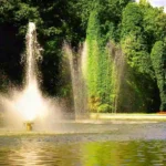 Outdoor water fountain spraying high jets of water in a pond with lush green trees in the background.