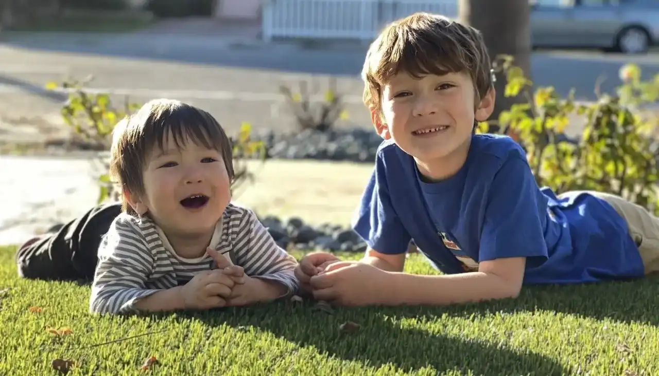 Two young children lying and smiling on artificial grass in a sunny front yard, showing how kid-friendly and safe synthetic turf can be.