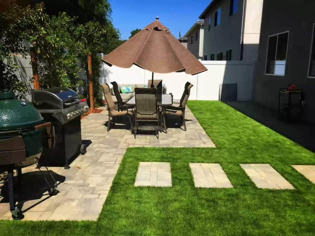 Modern backyard with artificial grass, patio dining set, paver walkway, and outdoor grill—showcasing clean, low-maintenance landscaping ideas.