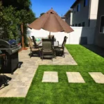 Modern backyard with artificial grass, patio dining set, paver walkway, and outdoor grill—showcasing clean, low-maintenance landscaping ideas.