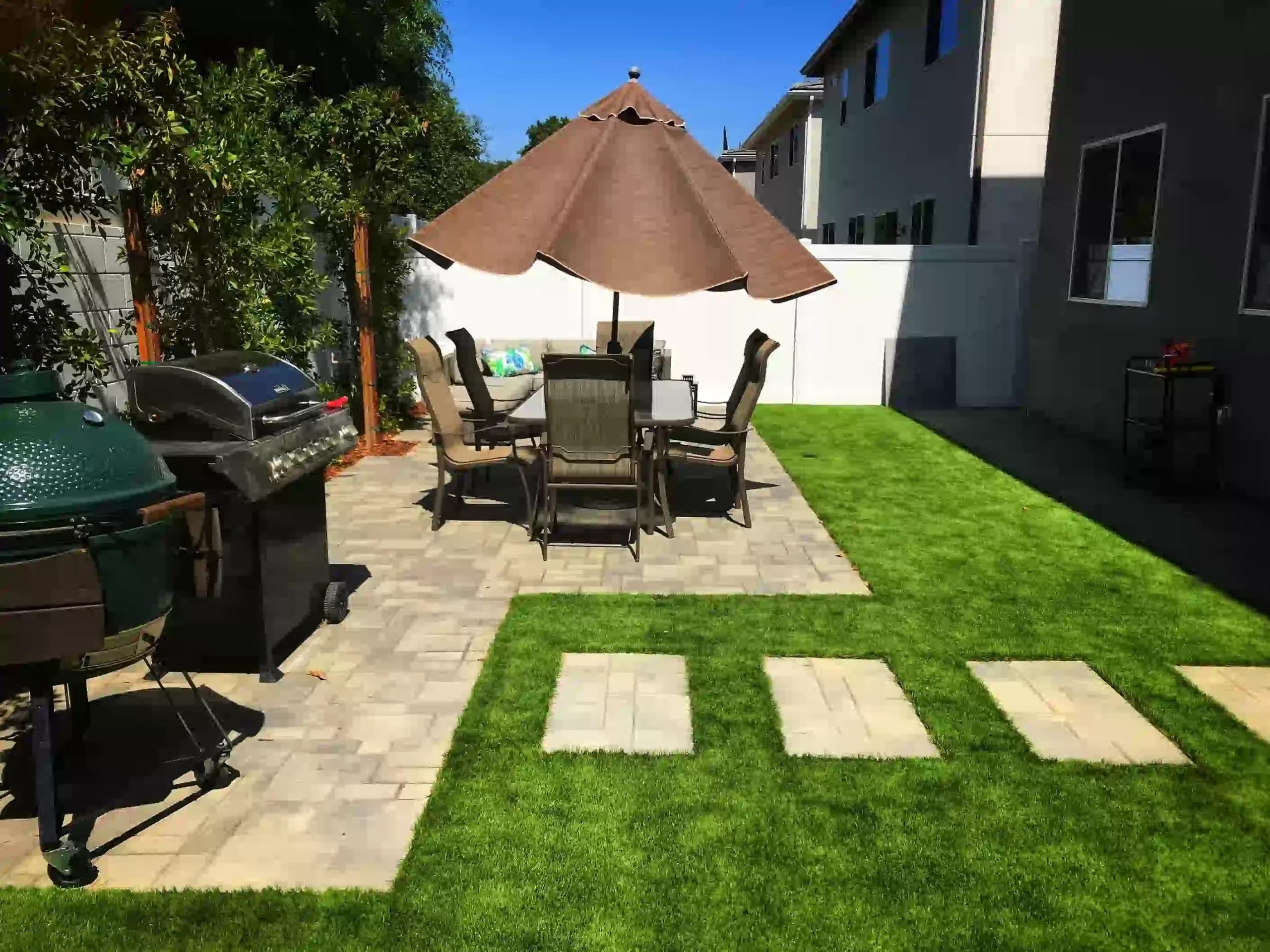Modern backyard with artificial grass, patio dining set, paver walkway, and outdoor grill—showcasing clean, low-maintenance landscaping ideas.